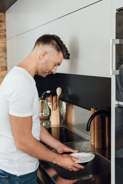 Schöner Mann hält nassen Teller in der Nähe von Wasserhahn und Spüle in Küche — Stockfoto