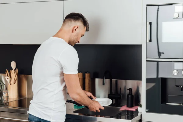 Schöner Mann steht und wäscht nassen Teller in Küche — Stockfoto