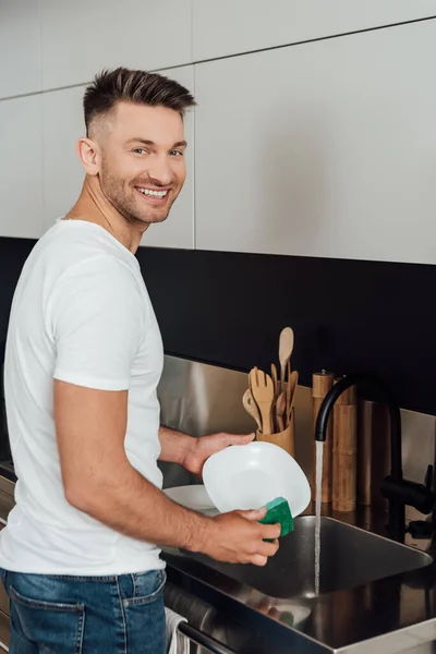 Hombre alegre sonriendo mientras sostiene la esponja y el plato blanco - foto de stock