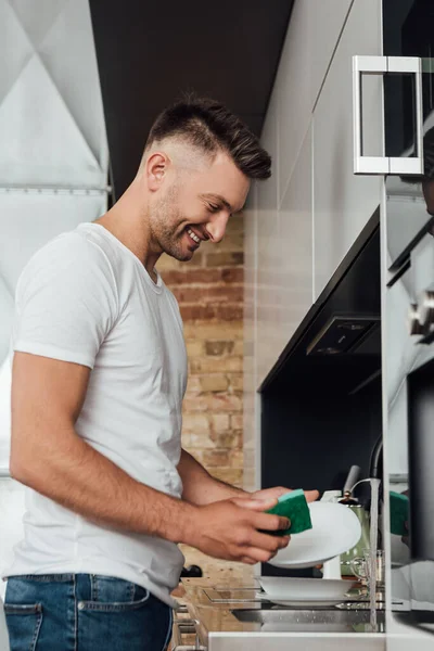 Feliz hombre sonriendo mientras sostiene esponja y plato blanco - foto de stock