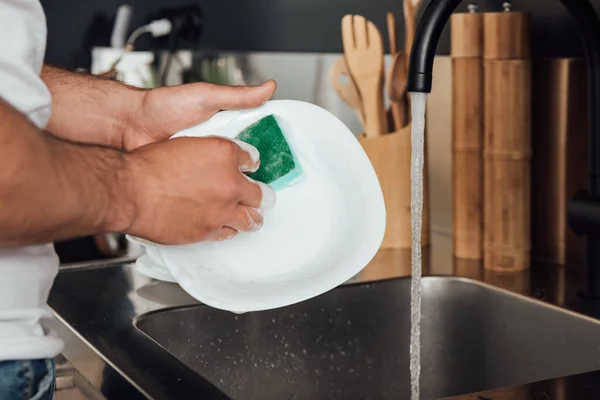 Vista recortada del hombre sosteniendo la esponja mientras lava la placa blanca en la cocina - foto de stock