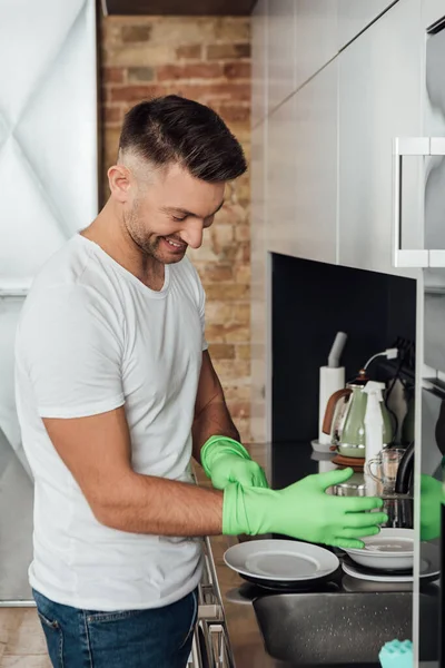 Hombre sonriente con guantes de goma cerca de platos blancos en la cocina - foto de stock