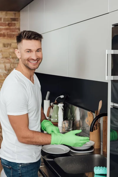 Uomo allegro indossare guanti di gomma vicino piatti bianchi in cucina — Foto stock