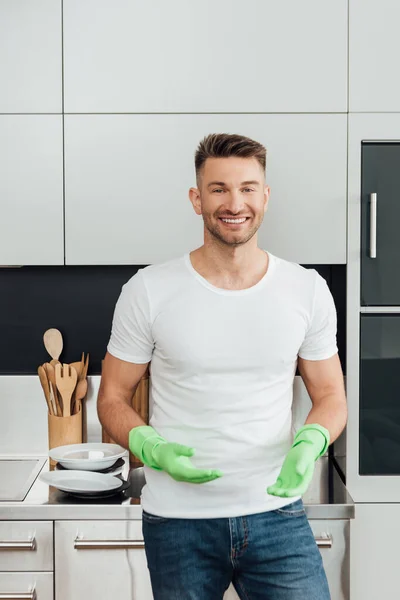 Uomo allegro in guanti di gomma in piedi vicino a piatti bianchi in cucina — Foto stock