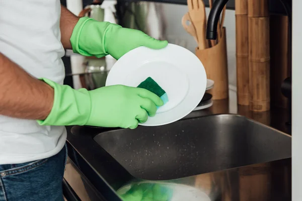 Обрезанный вид человека в резиновых перчатках, моющая тарелка с губкой — стоковое фото