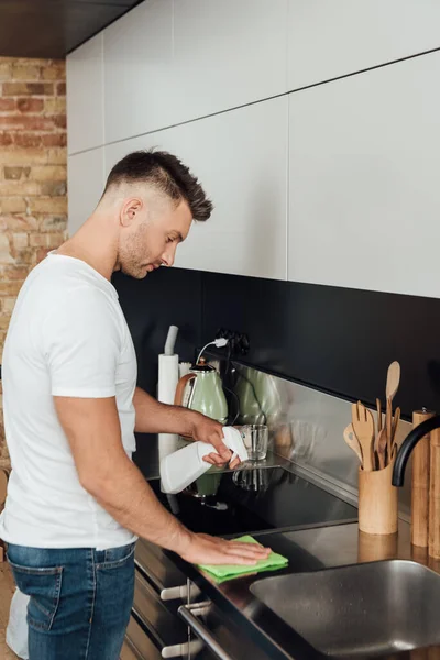 Homem bonito segurando pano e spray garrafa enquanto a mesa de limpeza — Fotografia de Stock