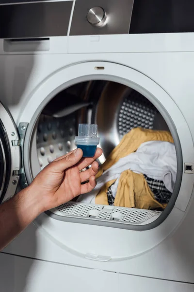 Обрезанный вид человека, держащего мерную чашку с голубым моющим средством возле стиральной машины с грязной одеждой — стоковое фото