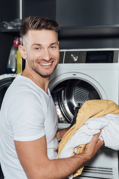 Homem feliz segurando roupa suja perto da máquina de lavar roupa — Fotografia de Stock