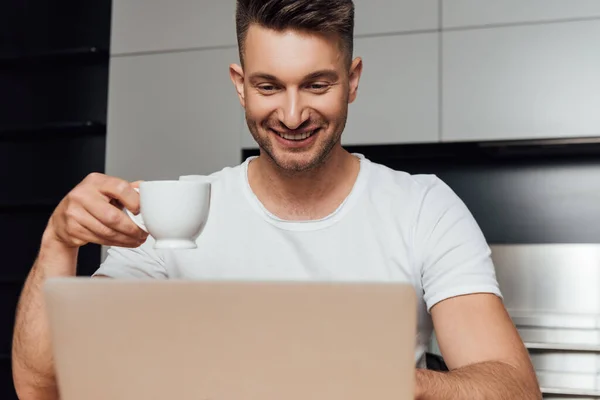 Selective focus of happy freelancer holding cup of coffee and looking at laptop — Stock Photo