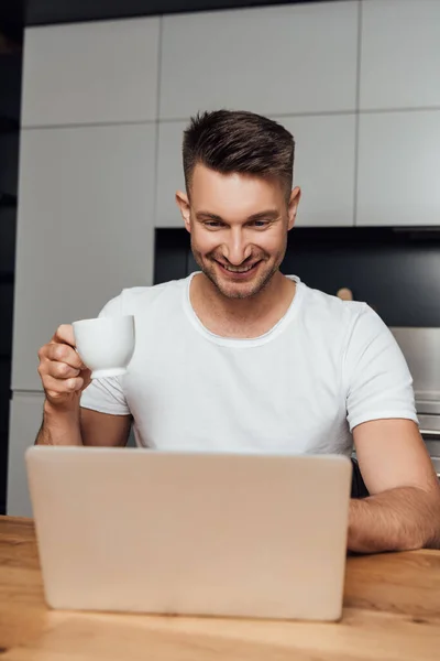 Cheerful freelancer holding cup of coffee and looking at laptop — Stock Photo