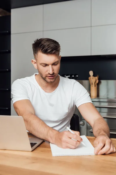 Konzentrierter Mann hält Stift in der Hand und schaut beim Online-Studium auf Notizbuch neben Laptop — Stockfoto