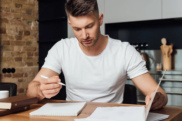 Handsome man holding pen and looking at notebook on table — Stock Photo