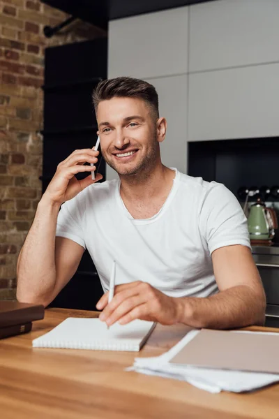 Foco seletivo de homem sorridente falando no smartphone perto de notebook e papelada na mesa em casa — Fotografia de Stock