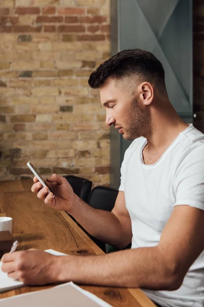 Side view of man using smartphone and writing on notebook at table in kitchen — Stock Photo