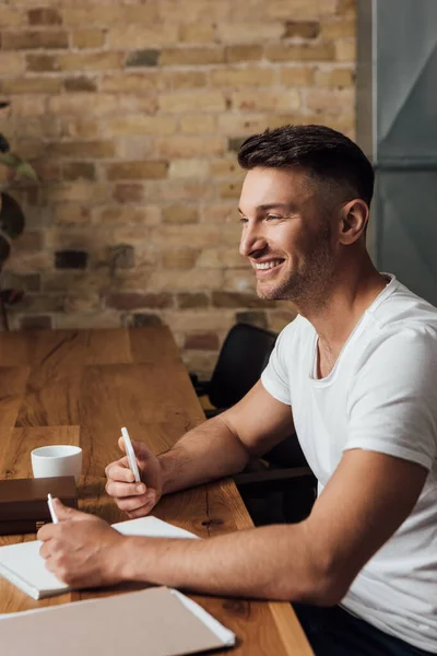 Вид сбоку улыбающегося человека, держащего смартфон рядом с бумагой и книгами на кухонном столе — стоковое фото