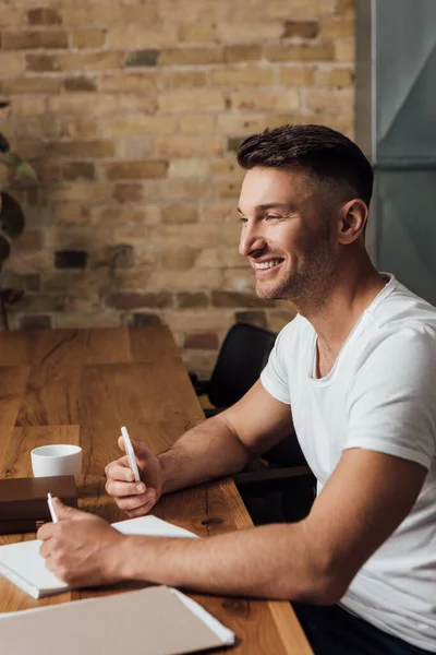 Vista lateral del hombre sonriente sosteniendo teléfono inteligente cerca de papeleo y libros en la mesa de la cocina - foto de stock