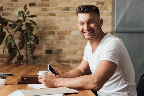 Vista lateral del hombre sonriendo a la cámara mientras sostiene el teléfono inteligente cerca de documentos, libros y taza en la mesa - foto de stock