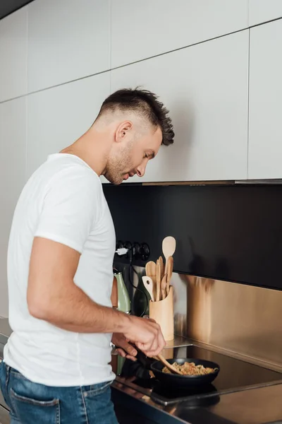 Side view of handsome man cooking noodles in frying pan in kitchen — Stock Photo