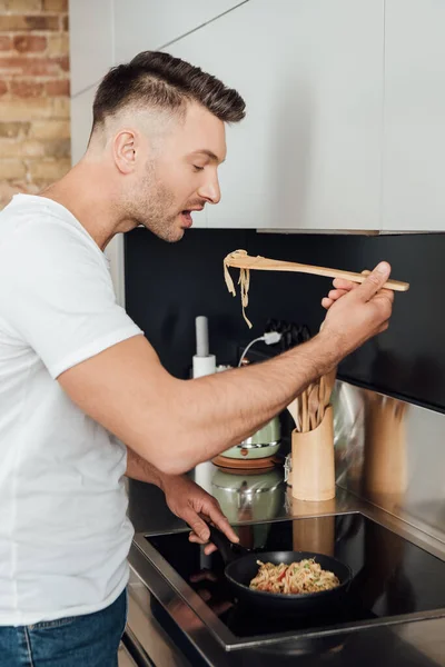 Vista lateral del hombre guapo sosteniendo espátula de madera mientras cocina fideos en la cocina - foto de stock