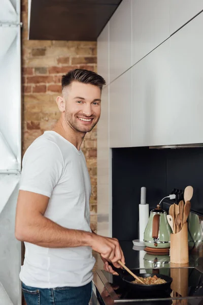 Vista lateral del hombre guapo sonriendo a la cámara mientras cocina fideos en la sartén en la cocina - foto de stock