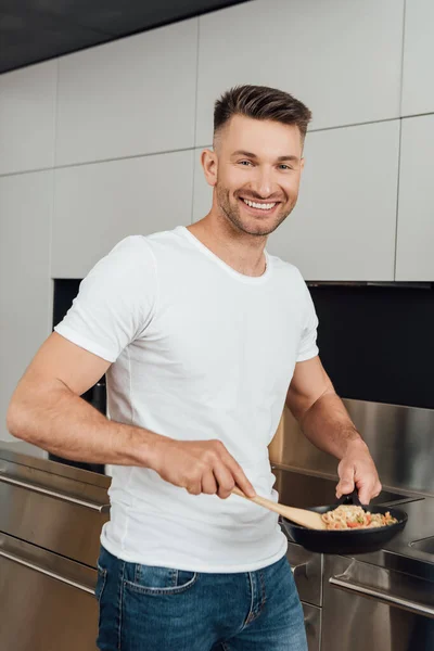 Handsome man smiling at camera while holding spatula and frying pan with noodles in kitchen — Stock Photo