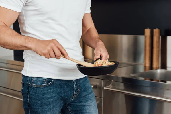 Vista recortada del hombre sosteniendo espátula de madera mientras mezcla fideos en la sartén en la cocina - foto de stock