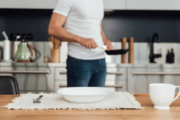 Selective focus of plate, cup and fork on table and man holding frying pan in kitchen — Stock Photo