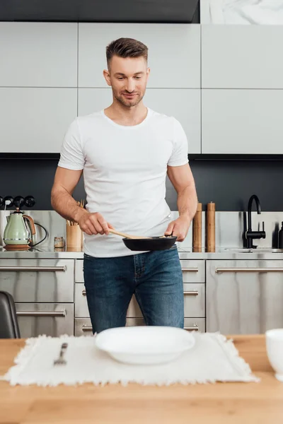 Selective focus of handsome man holding frying pan and spatula near plate and fork on kitchen table — Stock Photo