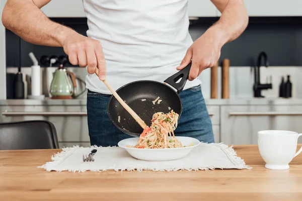 Cropped view of man pouring tasty noodles from frying pan in plate on kitchen table — Stock Photo