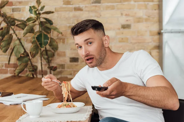 Man clicking channels while eating noodles near papers in kitchen — Stock Photo