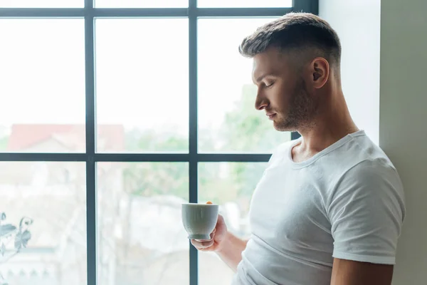 Seitenansicht eines gutaussehenden Mannes, der eine Tasse Kaffee hält, während er zu Hause am Fenster steht — Stockfoto