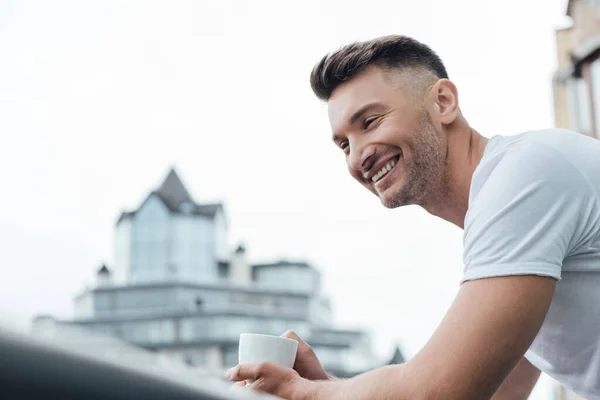 Selektiver Fokus eines gutaussehenden Mannes, der lächelt, während er eine Tasse Kaffee auf der heimischen Terrasse hält — Stockfoto