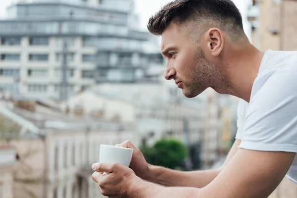 Vista lateral del hombre guapo sosteniendo la taza de café en el balcón - foto de stock