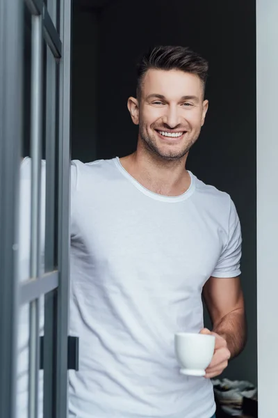 Selektiver Fokus eines gutaussehenden Mannes, der in die Kamera lächelt, während er zu Hause eine Tasse Kaffee in der Nähe der Tür hält — Stockfoto