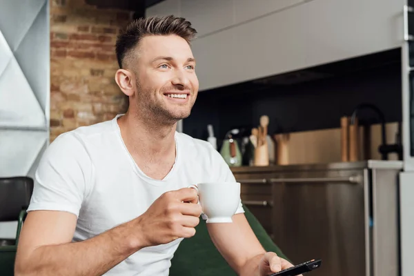 Sorrindo homem segurando xícara de café enquanto assiste tv no sofá — Fotografia de Stock
