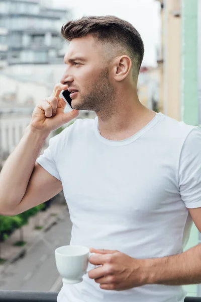 Hombre guapo hablando en el teléfono inteligente y sosteniendo la taza de café en la terraza - foto de stock
