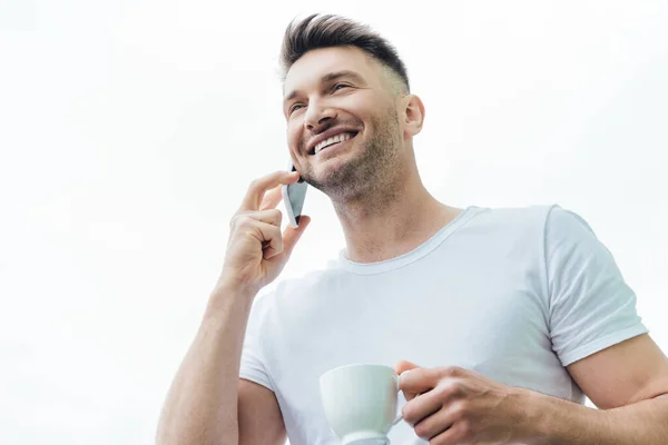 Low angle view of smiling man talking on smartphone and holding cup of coffee isolated on white — Stock Photo