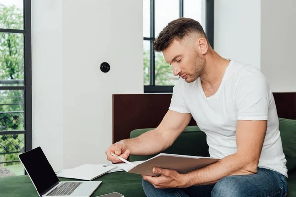 Guapo freelancer sosteniendo documentos y pluma cerca de gadgets en el sofá en casa - foto de stock