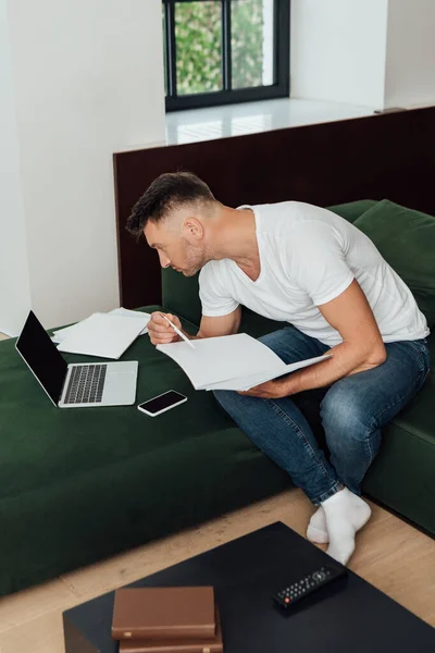 Freelancer sostiene portátil cerca de dispositivos digitales y libros en la sala de estar - foto de stock