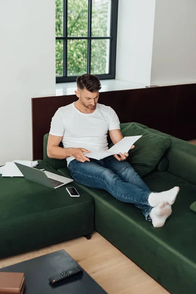 Selective focus of man holding notebook near digital devices and papers on couch — Stock Photo