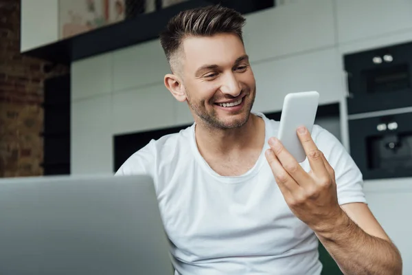 Выборочный фокус улыбающегося фрилансера, держащего смартфон возле ноутбука дома — стоковое фото