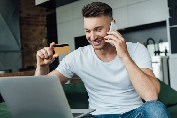 Selective focus of smiling man holding credit card while talking on smartphone near laptop in living room — Stock Photo