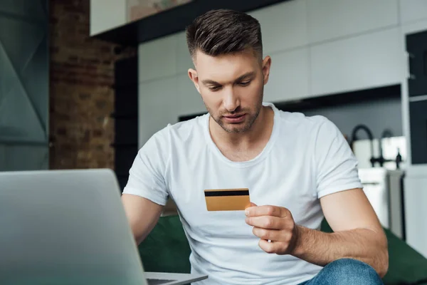 Foco seletivo do homem segurando cartão de crédito e usando laptop na sala de estar — Fotografia de Stock