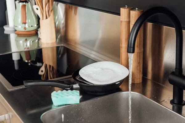 Selective focus of plate, frying pan and sponge in soap near kitchen sink with faucet — Stock Photo