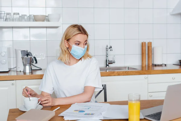 Freelancer in medical mask looking at laptop near drinks, glass of orange juice, charts and graphs — Stock Photo