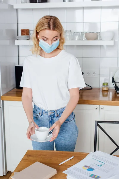 Mujer joven en máscara médica sosteniendo platillo y taza de café - foto de stock