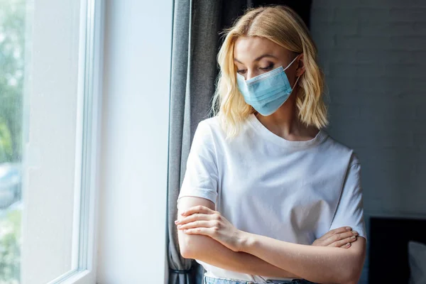 Giovane donna in maschera medica con braccia incrociate in piedi vicino alla finestra a casa — Foto stock