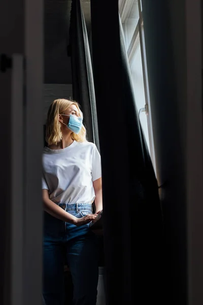Enfoque selectivo de la mujer joven en máscara médica de pie cerca de la ventana en casa - foto de stock