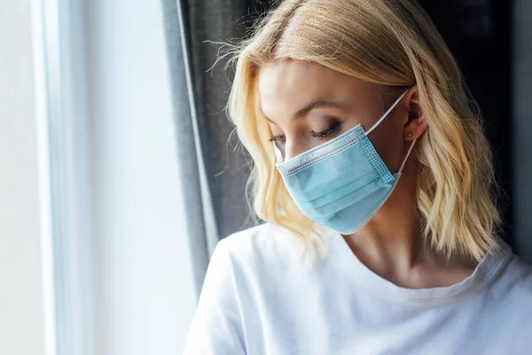 Donna in maschera medica guardando verso il basso vicino alla finestra a casa — Foto stock