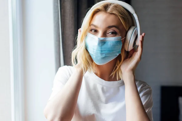 Mujer joven en máscara médica tocando auriculares inalámbricos en casa - foto de stock